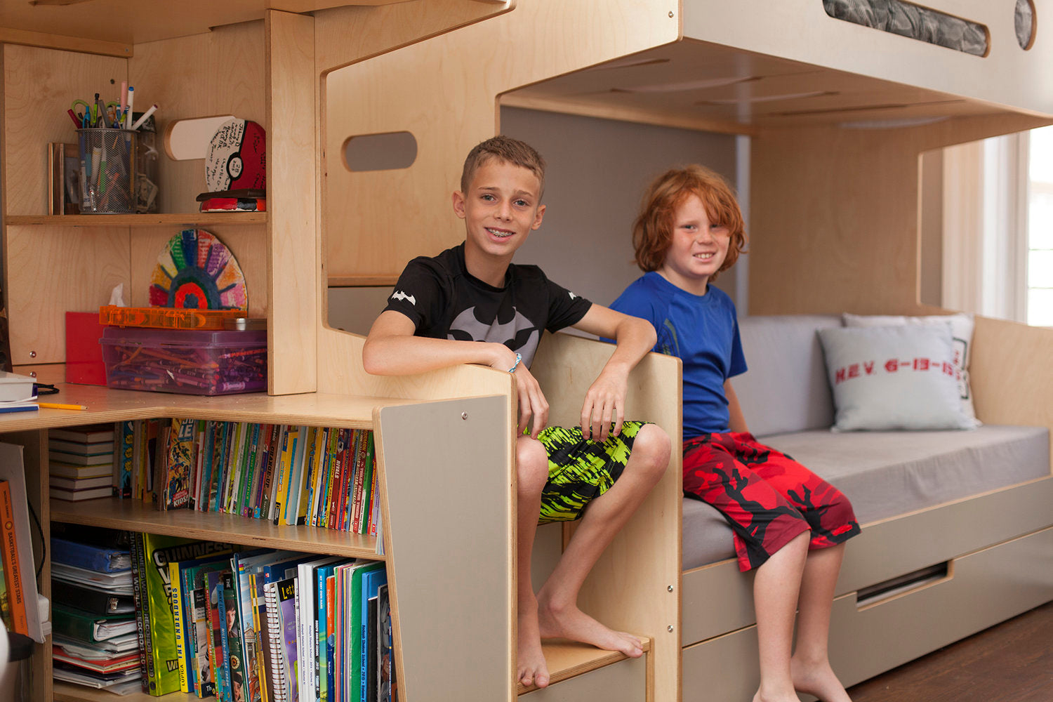 Two boys sitting in a bedroom with a loft bed and built-in desk.