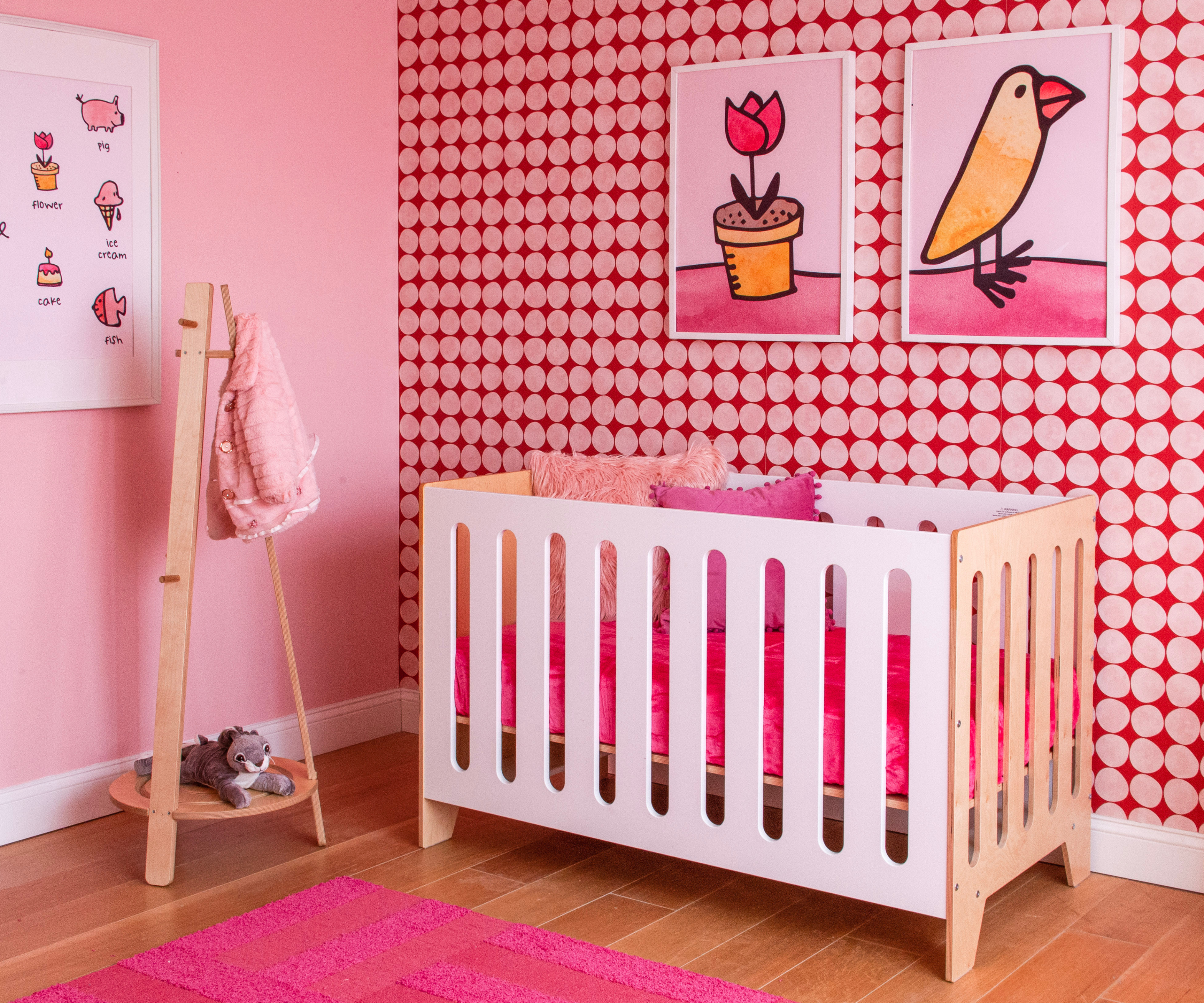 Nursery room with pink patterned wallpaper, white crib, and colorful artworks on the wall.