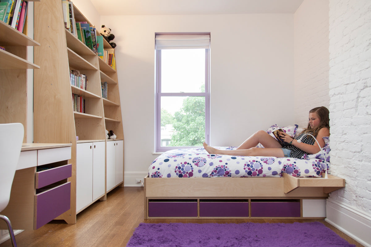 Modern bedroom, white wall, bookshelf, desk, bed with purple bedding, large window.