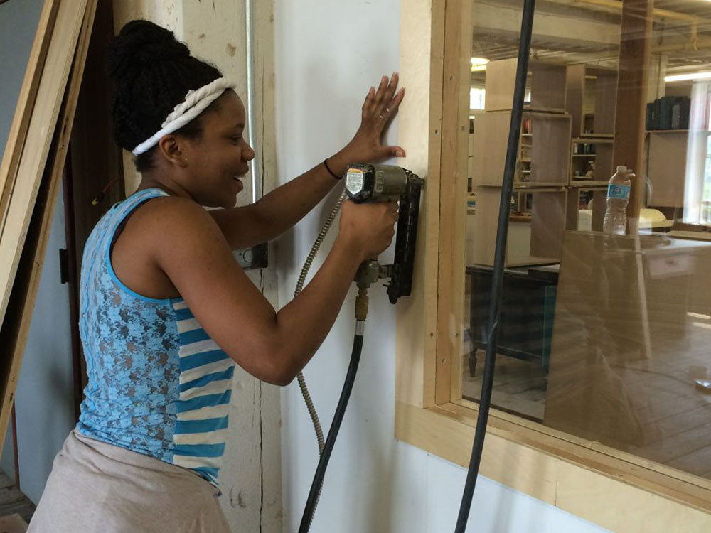 Woman in a striped tank top using a nail gun to secure a wooden frame in a workshop, focused on her work.