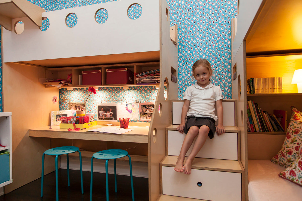 Young boy sitting on ladder of a loft bed above a study area with shelves and colorful wallpaper.