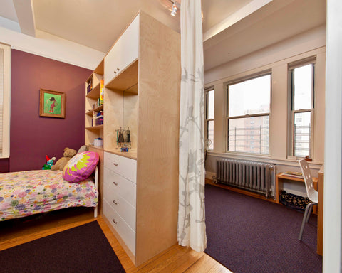 Cozy corner of a child's room with a bed, built-in shelving and desk, under soft purple walls and windows.