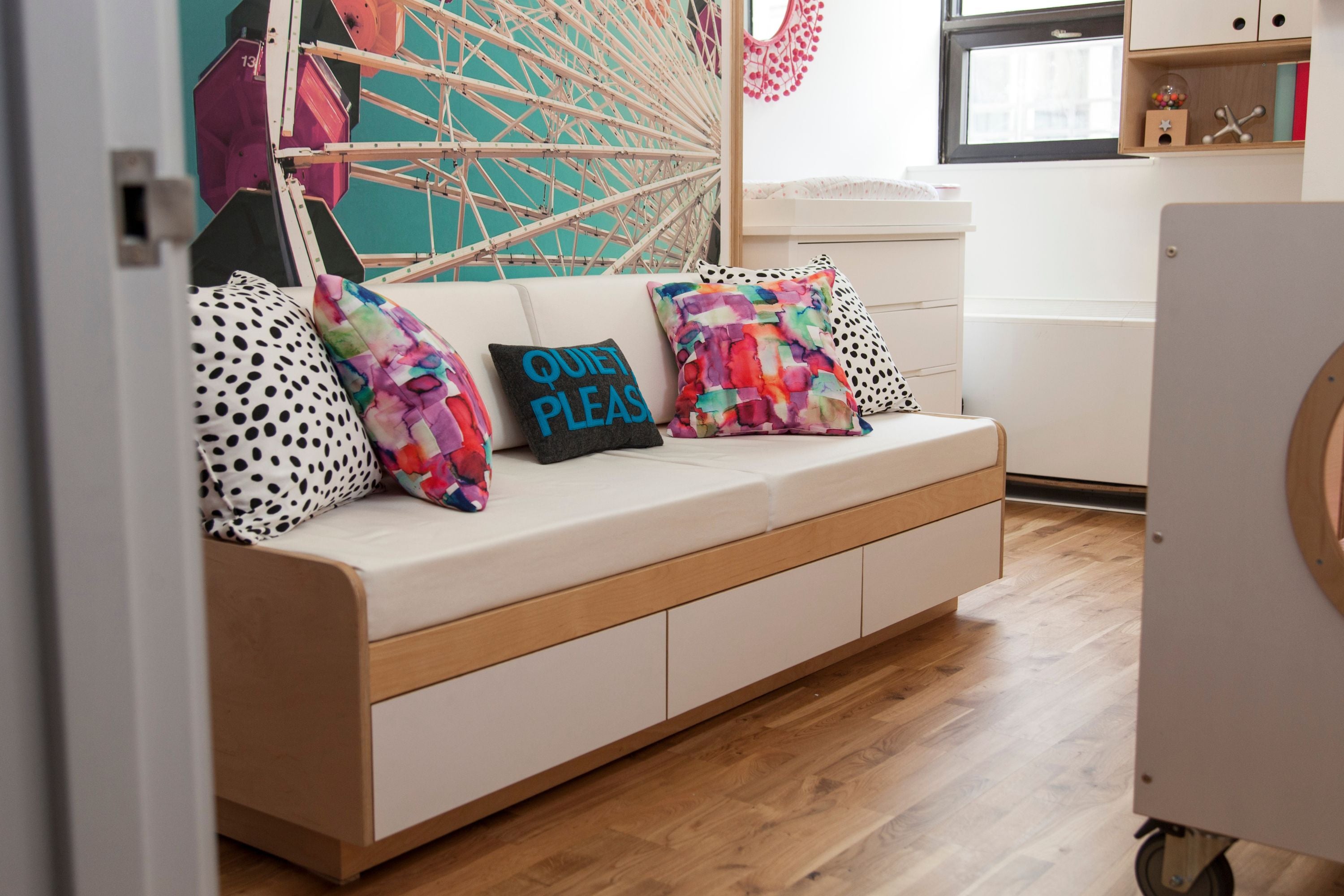 A modern full-size Murphy bed positioned above a built-in couch with white cushions and wooden frame. The couch features storage drawers underneath. The room has colorful accent pillows and a large Ferris wheel mural on the wall, creating a playful and functional design