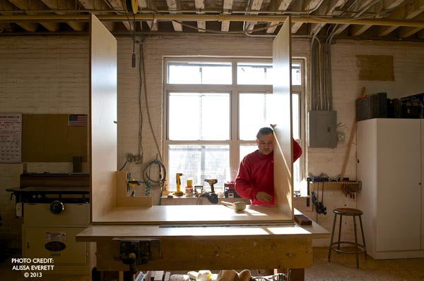 Person in red hoodie sands wood in a bright workshop, showcasing skilled woodworking.