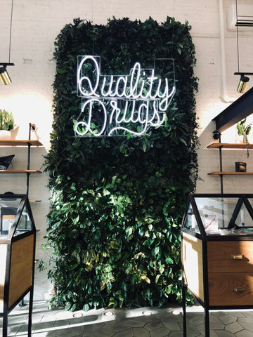 A green, leafy vine wall with a neon sign reading 'Quality Drugs' is in a cannabis dispensary