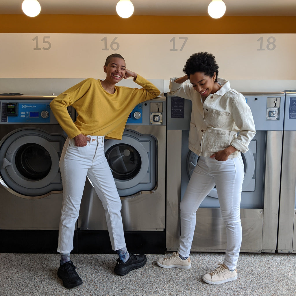 Corinna and Theresa Williams laugh and smile while perched next to sleek looking dryers, wearing white Madewell jeans