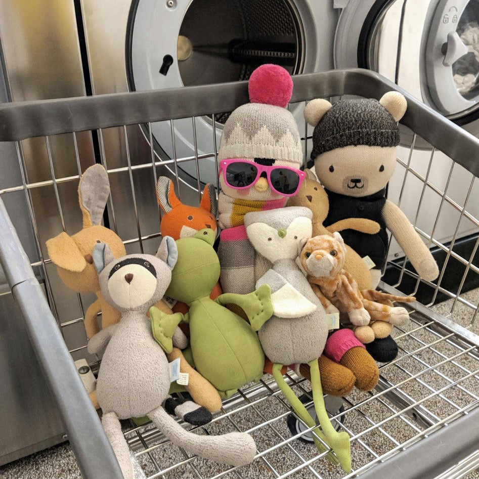 An adorable group of stuffed animals assembled in a laundry cart in front of an open washer