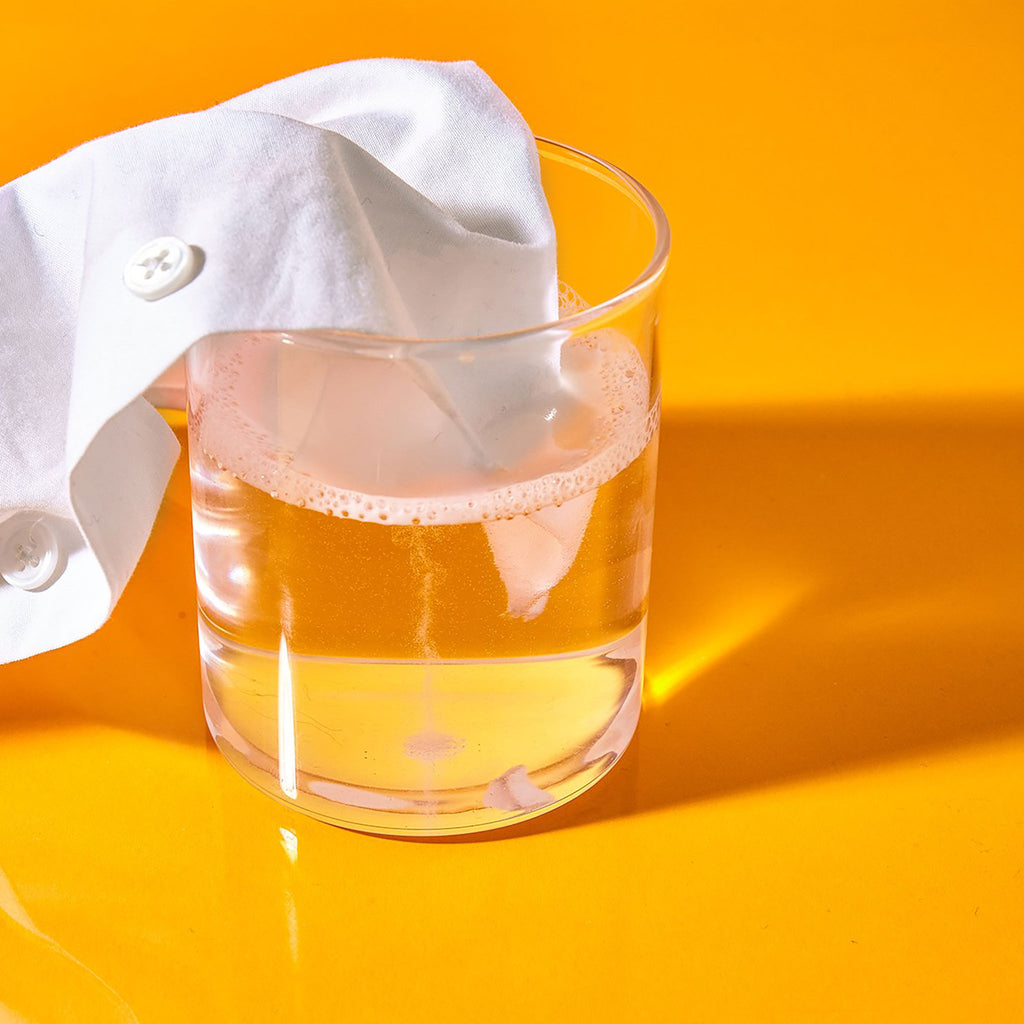A section of a stained white button down shirt is submerged in a clear glass of water with oxygen booster
