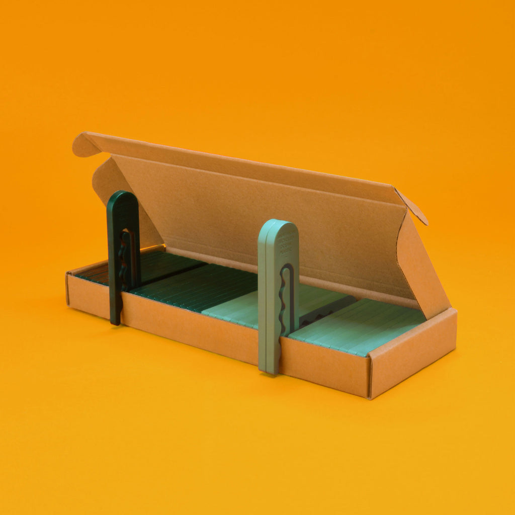 An open box of 40 recycled ocean plastic clothespins in two shades of green, displayed on an orange background
