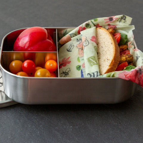 A sandwich wrapped in Bees Love These sits in a divided stainless steel lunch tin with cherry tomatoes and a nectarine.