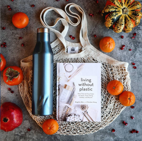 Living Without Plastic book cover on a cotton grocery bag with orange fruits and a blue metal reusable water bottle