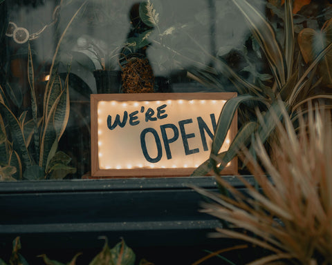 A sign in a window partially obscured by plants that reads We're Open