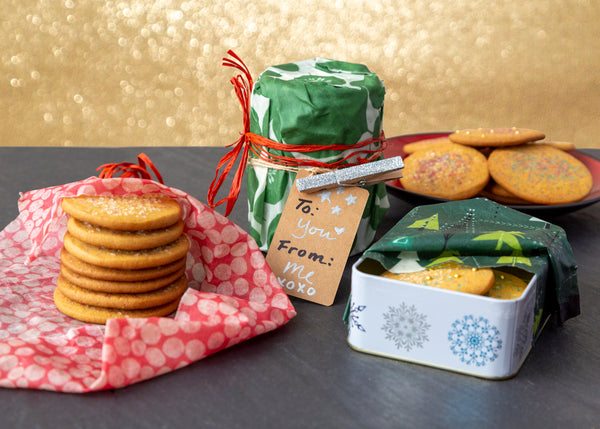 Sugar cookies being prepped for gifting in front of a sparkly gold background. left to right, a stack of cookies on a dot-print wrap, a wrapped cylinder of cookies in a leafy green wrap with a red raffia ribbon and "from me to you" gift tag, a plate of cookies in the background, and a snowflake-print tin of cookies with a winter trees wrap.