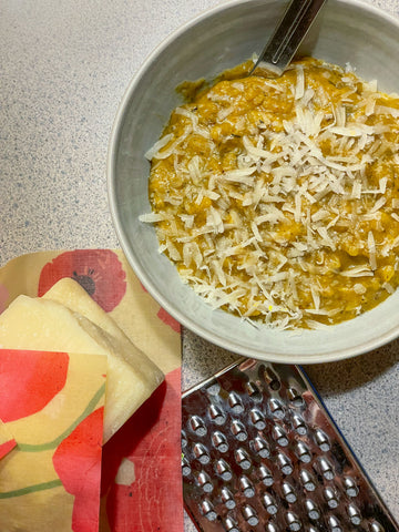 A bowl of pumpkin risotto on a home kitchen counter, with a Painted Poppy Z Wrap and a hunk of cheese and a grater beside it