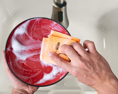 Hands with an orange Skoy Swedish Dishcloth wash a soapy red plate over a white sink.