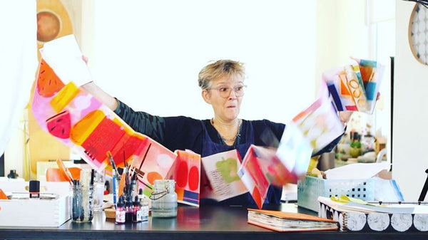 Susy Pilgrim sitting at her studio desk in her light and bright studio, excitedly holding up a bunting or garland of colorful squares