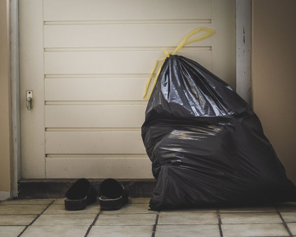A black plastic trash bag, full, sits ominously waiting to be taken out next to a door and a pair of shoes.