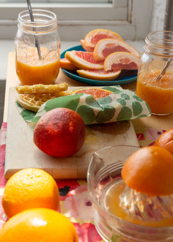 Assorted citrus slices and fresh squeezed juice; a Painted Poppy Z Wrap covers the counter, with a small Z Wraps beeswax food wrap covering half a citrus left on the cutting board 