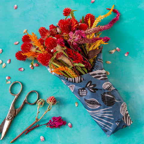 a bouquet of red and yellow flowers is wrapped in a Z Wraps beeswax wrap on a teal background next to some old scissors and a couple stems