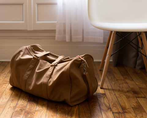 A tan duffle sits on the floor next to a white chair, ready for the next trip