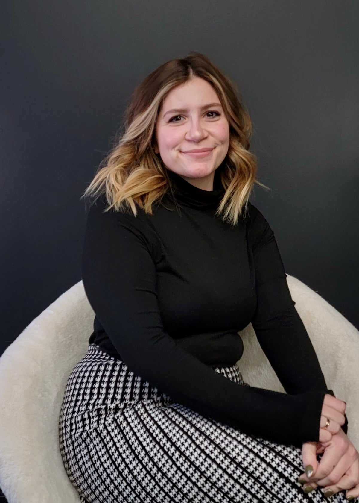 Lady with mid length hair and blonde highlights. Long sleeve black shirt and a white and black skirt. Sitting in a white chair against a black background.