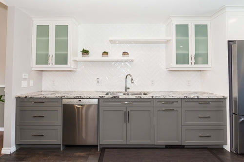 Grey lower cabinets with white upper cabinets on a grey and brown vinyl floor. White and grey granite countertops, white rectangular herringbone tile, white floating shelves.