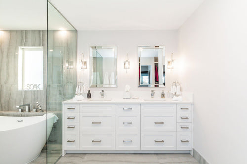 White vanity cabinets, a white quartz countertop, and a mirror framed mirror. Grey travertine tile, chrome plumbing fixtures, and a white freestanding tub.