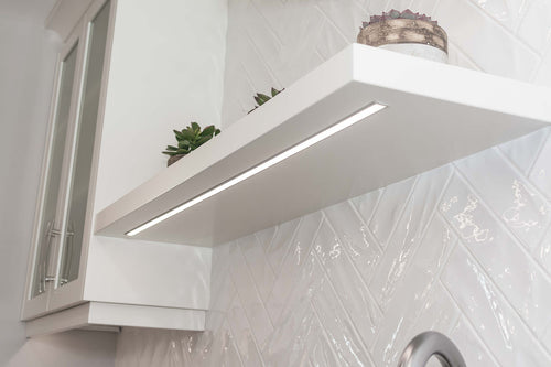 White upper cabinets with clear glass inserts, white floating shelves with under cabinet lighting, white rectangle herringbone tile.