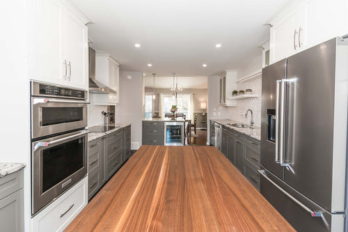 Grey lower cabinets with white upper cabinets on a grey and brown vinyl floor. White and grey granite countertops, white rectangular herringbone tile, white floating shelves, and a grey island with a walnut countertop..