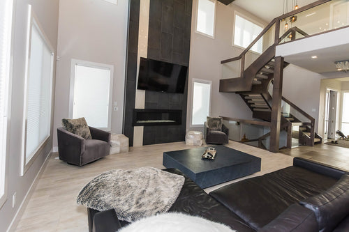 Light grey floor tile, black and white floor to ceiling fireplace tile, linear fireplace, black sectional couch, grey side chairs, grey stairwell with clear glass railing.