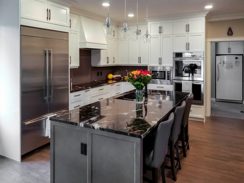 White perimeter cabinets with light grey island cabinet. Dark granite countertop. 3 grey stools at island with vase of flowers. 4 clear glass pendants over island.