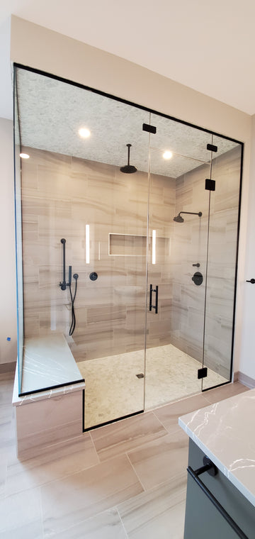 Large steam shower with light grey wall tile, grey quartz bench top, black plumbing fixtures, clear glass, and white pebble shower floor and ceiling.