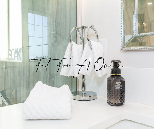 Freestanding hand towel hanger with white towels, a white quartz countertop, and soap. A mirror framed mirror with grey travertine tile.