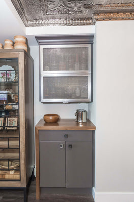 Tin ceiling, light grey walls, dark grey cabinet with wood countertop and mesh upper cabinet doors.