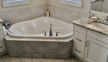 Corner built in tub with white cabinets, granite countertops, and beige square tiled floor and tub surround.