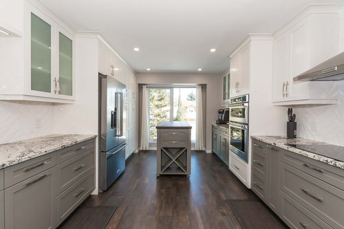 Grey lower cabinets with white upper cabinets on a grey and brown vinyl floor. White and grey granite countertops, white rectangular herringbone tile, white floating shelves, and a grey island with a walnut countertop..