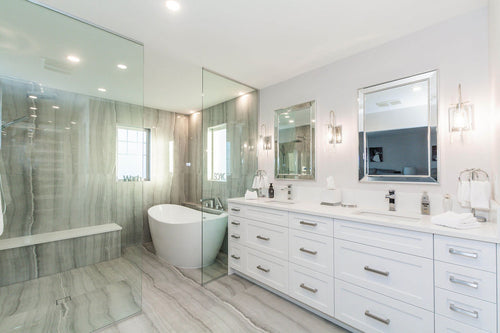 White vanity cabinets, a white quartz countertop, and a mirror framed mirror. Grey travertine tile, chrome plumbing fixtures, and a white freestanding tub.