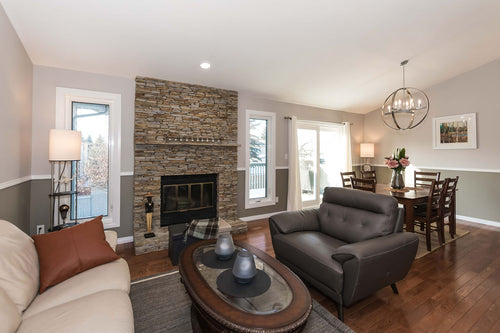 Brown hardwood floor with grey oversized chair. The living room is open to the living room. Natural grey and beige stacked stone with a wood burning fireplace. The walls have a chair rail with grey below the chair rail and light gray above the chair rail.