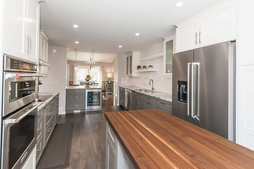 Grey lower cabinets with white upper cabinets on a grey and brown vinyl floor. White and grey granite countertops, white rectangular herringbone tile, white floating shelves, and a grey island with a walnut countertop.