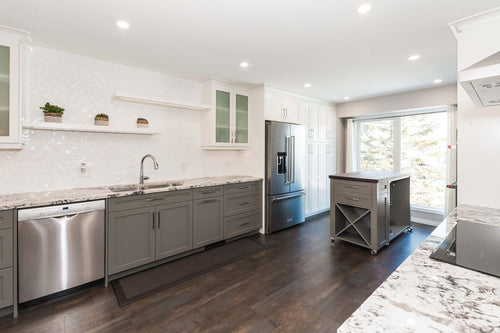 Grey lower cabinets with white upper cabinets on a grey and brown vinyl floor. White and grey granite countertops, white rectangular herringbone tile, white floating shelves, and a grey island with a walnut countertop.