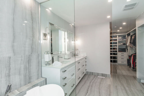 White vanity cabinets, a white quartz countertop, and a mirror framed mirror. Grey travertine tile, chrome plumbing fixtures, and a white freestanding tub.