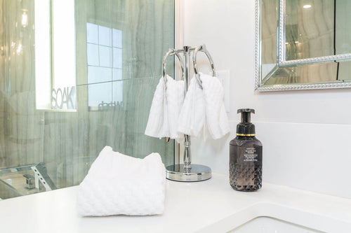 Freestanding hand towel hanger with white towels, a white quartz countertop, and soap. A mirror framed mirror with grey travertine tile.