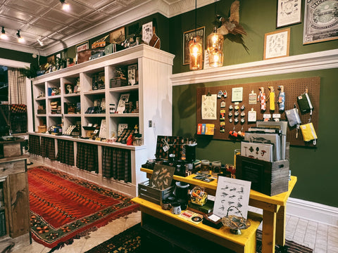 A warmly lit shop interior with dark green walls, bright rugs, and shelves full of artwork and gifts.