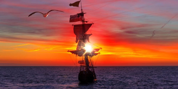 A tall sailed ship sails into a dramatic setting sun.