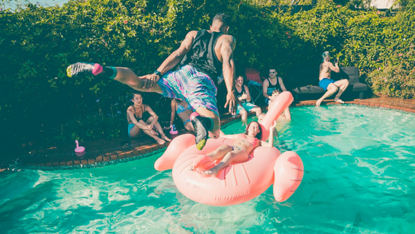 A group of people enjoying a casual pool party. A woman relaxes on a large inflatable flamingo pool float and her friend jumps in on her.