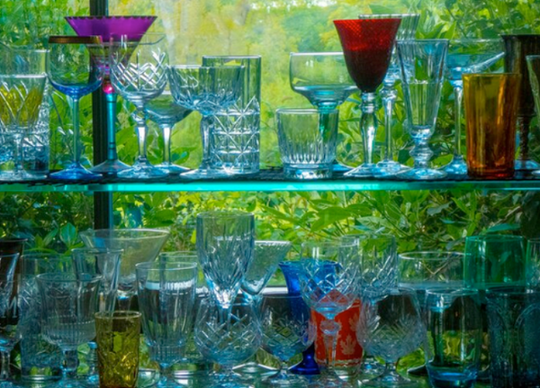 A collection of vibrantly colored glassware on a glass shelf in front of a window looking out into lush green scenery.