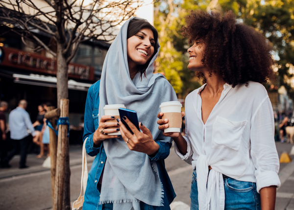 Two female friends enjoy a coffee-to-go while walking down a city street together.