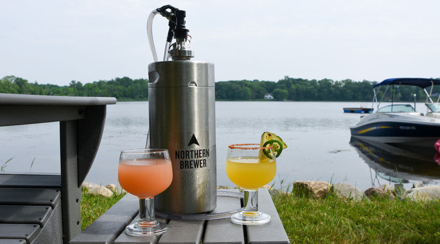 1-gallon keg and two cocktails on table next to lake