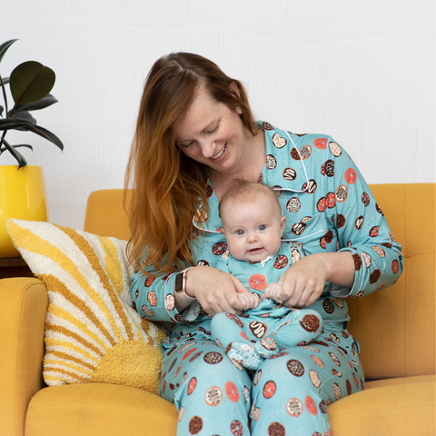 Mom is holding her baby in her lap while they sit on a yellow couch. The two of them are wearing matching blue pajamas with colorful donuts all over them. 