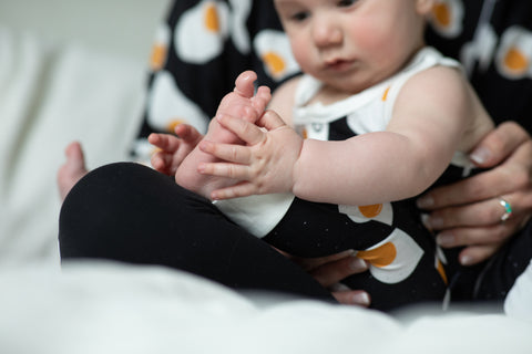 A baby plays with his foot while sitting in moms lap. They are wearing matching egg pajamas